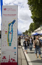 The Paris Plage urban beach. People strolling beside the River Seine along the Voie Georges Pompidou usually a busy road now closed to traffic opposite the Ile de la Cite with a map showing the attrac...