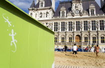 The Paris Plage urban beach. Young people playing beach volleyball behind a green beach hut with geckos painted on it in front of the Hotel de Ville Town Hall