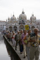 Aqua Alta High Water flooding in St Marks Square A party of tourists walking on an elevated walkway above the water with St Marks Basilica at the far end of the piazza