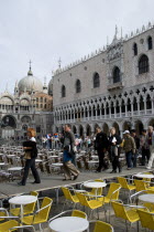 Aqua Alta High Water flooding in St Marks Square with tourists walking on an elevated walkway through restaurant tables by the Doges Palace with St Marks Basilica beyond