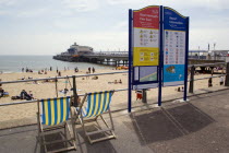 Deckchairs beside an information sign on The East Beach promenade with Bournemouth Pier beyond tourists on the sandy beach and paddling in the sea.