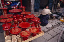 Red lacquered dishes and pots displayed on pavement  with two women sitting at side.