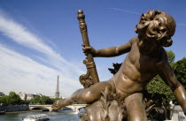 Bronze statue of a cherub on the Pont Alexandre III bridge over the River Seine with a Bateaux mouches pleasure boat with tourists passing below and the Eiffel Tower in the distanceFrench Western Eur...