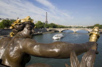 Bronze statue of a woman holding a flame on the Pont Alexandre III bridge over the River Seine with a Bateaux mouches pleasure boat with tourists passing below and the Eiffel Tower in the distanceFre...