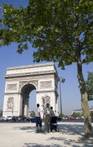 Tourist family standing in the shade of a tree in Place de Charles de Gaulle looking at the Arc de Triomphe with traffic passing around itFrench Western Europe European