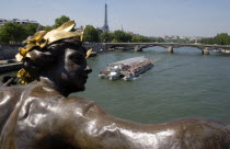 A bateaux mouches pleasure boat with sightseeing tourists passes below a bronze statue of a woman on the Pont Alexandre III bridge across the River Seine with the Eiffel Tower in the distanceEuropean...