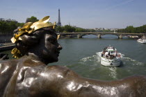 A boat with sightseeing tourists passes below a bronze statue of a woman on the Pont Alexandre III bridge across the River Seine with the Eiffel Tower in the distanceEuropean French Western Europe