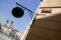 The Old Town Square. Art gallery and souvenir shop sign hanging on a wall corner below a street sign with the Cgurch of St Nicholas on the far side of the squarePraha Ceska Eastern Europe European Re...