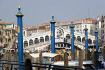 The Rialto Bridge with tourist crowds spanning the Grand Canal seen through wooden pillars for mooring boats by the side of the canal