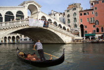 A gondola with sightseeing tourists passing beneath the Rialto Bridge spanning the Grand Canal lined with people at cafe tables under umbrellas