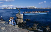 Family posing for photograph beside the Little Mermaid statue with passing tourist boat behind and city skyline beyond. Danish Danmark European Kids Northern Europe Scandinavia