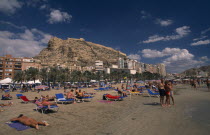 Alicante.  Sunbathers on sandy beach lined with palm trees and apartment buildings overlooked by castle on top of steep  rocky hillside behind.Beaches Espainia Espana Espanha Espanya European Hispani...