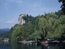 Tourist boats on Lake Bled with castle partly seen amongst trees high above.  Few people on lake shore below.European Scenic Slovenian Slovenija Southern Europe