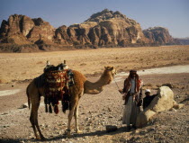 Bedouin man and child standing beside camel carrying decorated  woven saddle and panniers in desert area near Wadi Rum.Al-Urdunn Asian Children Jordanian Kids Male Men Guy Middle East Scenic Male Man...