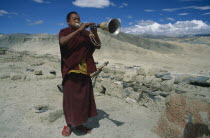 Tibetan Buddhist monk blowing long  decorated horn on monastery roof to summon the faithful to prayer.  Barren  mountainous landscape behind.Asia Asian Nepalese Religion Religion Religious Buddhism B...