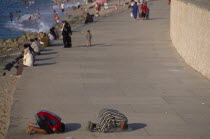 Corniche Waterfront . People sitting on the edge of the promenade facing the sea with two men knelling on the ground taking part in evening prayers in the foreground.Worshipping. Prayer African Middl...