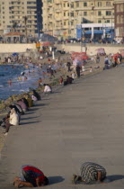 Corniche Waterfront . People sitting on the edge of the promenade facing the sea with two men knelling on the ground taking part in evening prayers in the foreground.WorshipAfrican Middle East North...