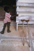 Child with both legs in braces sitting in open doorway of polio unit beside pair of crutches leaning against outside wall.African Eastern Africa Kids Tanzanian Children