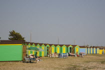 Green and yellow beach huts on shingle beach with sunbathers on sun loungers