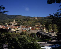 Prats-de-Mollo-la-Preste.  Mountain town with typical painted houses with red tiled rooftops and Fort Lagarde on hillside behind.  Road and tree tops in the foreground.