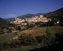 Mosset. View towards hillside village from the east with narrow houses with painted window shutters and red tiled rooftops. Backdrop of rocky hills scatteres with trees.