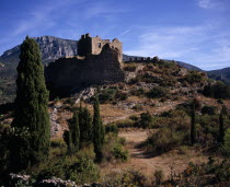 Chateau Padern.  Ruined fortifications perched on steep  rocky hillside above village amongst cypress trees with mountain backdrop.