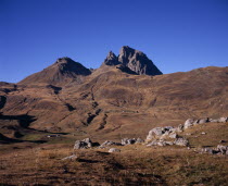 Pic du Midi d Ossau  2884 m  in Autumn. Eroded mountain-side and scattered houses with narrow winding road across centre-ground. Rocks in foreground.