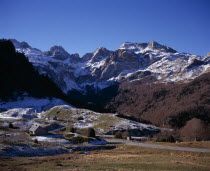 Winter view west from village of Peyrenere near mountain pass of Col du Somport towards Sierra de Bernera in light snow with two stone houses and road in foreground.