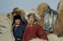 Bigersum negdel collective  Khalkha father and son in fleece-lined winter traditional silk tunics  standing with bactrian camel with rope bridle and leather saddle positioned between the two humps.Ea...