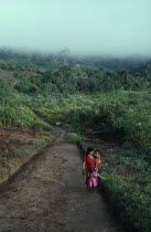 Makuna mother carrying baby and with young daughter in front of her walking between malocas along unmade road through cleared and cultivated rainforest.Tukano  Makuna Indian North Western Amazonia fa...