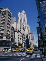 View along 5th Avenue in-between tall buildings with yellow taxi cabs on road and people walking on side walk