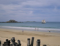 Le Sillon Beach and Fort National. People walking along golden sand with a sail boat on seen on water