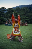 Figure in mask and costume performing Genroku  a bugaku dance originating in India and coming to Japan via China in the eigth century.  Depicts a snake-eater hunting for his food.  Line of musicians b...