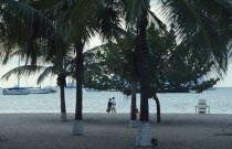 Couple on beach looking out over water and moored yachts framed by palms and other trees in foreground.