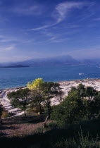 Sirmione.  View across Lake Garda from Villa Romana.  Flat outcrop of rock and trees in foreground with people sunbathing.  Distant mountains beyond.