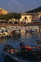 Garda.  Harbour view with cafe tables set out on quayside overlooked by pastel coloured buildings and with moored boats in the foreground.