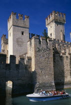 Sirmione.  Part view of crenellated walls and towers of the Rocca Scaligera medieval castle with passing tourist boat in foreground.