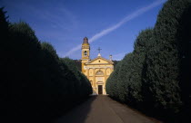 Yellow and white exterior facade of small church with bell / clock tower at side with tree lined road leading to entrance.