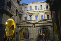 Street scene and Roman gateway at night with group of people framed in archway.  Light from street lamps and shop windows and people at restaurant tables on left.