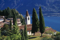 Pieve.  View of tiled rooftops  church and bell tower of village overlooking Western shore of Lake Garda.  Cypress trees in foreground and mountain backdrop.