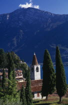 Pieve.  View of tiled rooftops  church and bell tower of village overlooking Western shore of Lake Garda.  Cypress trees in foreground and mountain backdrop.