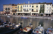 Bardolino. Harbour scene with people at outside tables of waterside cafes and line of moored boats in foreground.  Pastel coloured building facades reflected in the water.