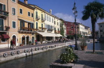 Bardolino.  Harbour and line of waterside bars and cafes painted in pastel colours and with awning pulled out over outside tables.  Moored boats in foreground.