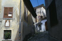 Narrow cobbled street with fresco decoration and plaster relief of the Virgin and Child on the exterior walls of the buildings and drinking fountain at top.