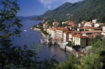 Cannero Riviera.  Waterside buildings with red tiled rooftops at foot of densely wooded hillside with view across lake part framed by tree branches.