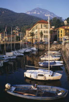 Menaggio.  Harbour with moored boats and waterside buildings with tree covered mountain backdrop.