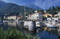 Menaggio.  Harbour with moored boats and waterside buildings with tree covered mountain backdrop.