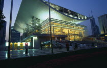 Opera House  Renmin Square.  Glass fronted exterior facade and upwardly curving roof illuminated at night with people standing or sitting in courtyard outside.