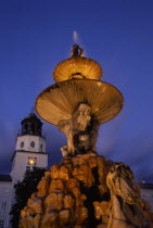 Part view of Baroque fountain with statues of horse and contorted figures and the Glockenspiel Carillon tower behind at night.