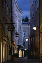 Narrow street of Judengasse at night lined by tall  grey  pink and cream painted buildings with street lamps extending from walls on metal brackets.